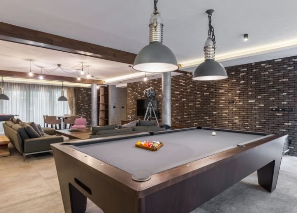 Interior view of a modern recreational room featuring a billiard table with racked balls, comfortable sofas, exposed brick walls, and industrial-style pendant lighting, suggesting a luxurious space for leisure and entertainment.