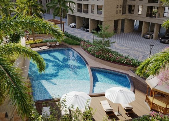 Aerial view of a luxurious swimming pool area within a premium residential complex. The image shows a large, irregularly shaped pool with clear blue water, surrounded by decking with lounge chairs under white umbrellas, lush green landscaping with palm trees, and a smaller adjacent pool or jacuzzi.