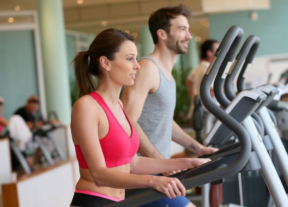 Two individuals exercising on elliptical machines in a modern gym. The woman in the foreground wears a pink sports bra, while the man behind her wears a grey tank top. Other gym-goers and equipment are visible in the background, suggesting a well-equipped fitness facility.