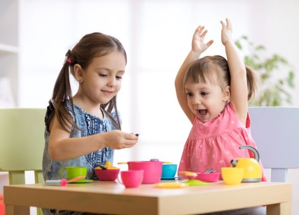 Two young girls engaged in imaginative play with toy kitchen utensils at a small table. One girl looks intently at something in her hands, while the other raises her arms with an expression of joy.